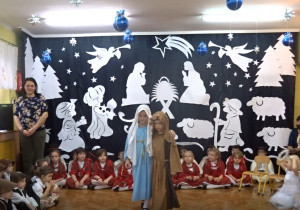 Na tle bożonarodzeniowej dekoracji stoją dzieci w strojach Maryji i Józefa.